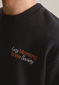 Felpa nera con colletto a coste, caratterizzata da un ricamo con la scritta "Società della Colazione Pigra" in bianco e arancione. Tessuto morbido.