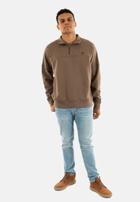 Felpa marrone con zip e collo alto, abbinata a jeans azzurri e stivali marroni. La felpa presenta un piccolo logo sul petto.