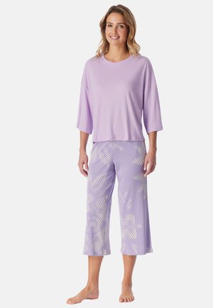 Femme debout pieds nus portant un haut ample lilas et un pantalon culotte à jambes larges avec des motifs géométriques légers sur fond blanc.