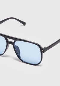 Schwarze Sonnenbrille mit Metallgestell, rechteckigen hellblau getönten Gläsern und einem Doppelsteg vor schlichtem hellem Hintergrund.