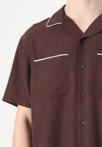 Braunes Kurzarmhemd mit einem Reverskragen, zwei Brusttaschen und kontrastierenden weißen Pipings entlang des Kragens und der Taschen.