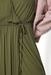 Olive green dress with a cinched waist and gathered fabric. A soft pink cardigan is partially visible on the side, featuring a textured finish.