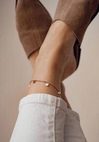 Braccialetto alla caviglia in oro rosa con piccoli charm circolari, indossato su una caviglia nuda. Scarpa tacco in camoscio marrone e risvolto in denim bianco visibili.