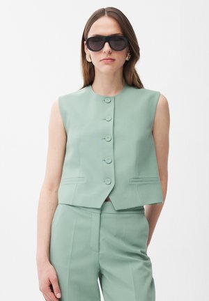 Frau trägt ärmellose, mintgrüne Weste mit Knöpfen, passende Hose, große schwarze Sonnenbrille und goldene Ohrringe vor weißem Hintergrund.
