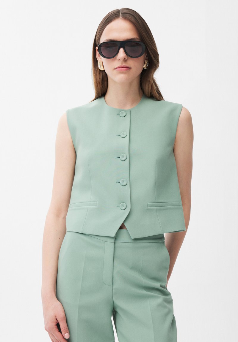 Donna con gilet senza maniche verde menta abbottonato, pantaloni coordinati, grandi occhiali da sole neri e orecchini d'oro su sfondo bianco.