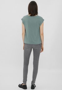 Grünes Kurzarmoberteil mit lockerer Passform, kombiniert mit figurbetonten grauen Hosen. Das Outfit wird mit schwarzen flachen Schuhen abgerundet.