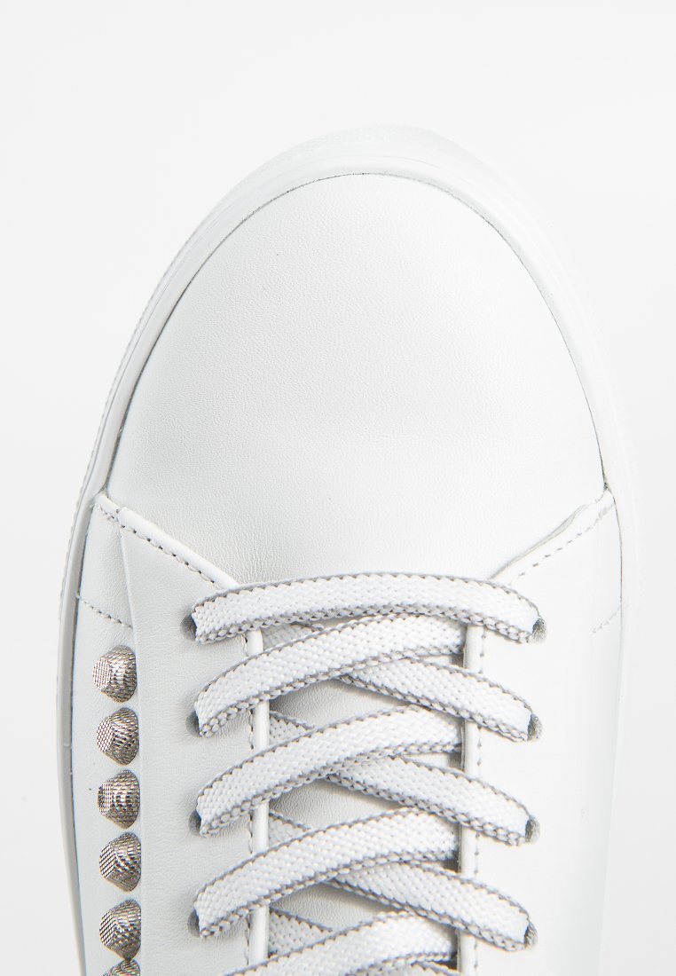 Nahaufnahme eines weißen Sneakers mit weißen Schnürsenkeln und metallischen Nieten entlang der Seite, von oben auf einem weißen Hintergrund gezeigt.
