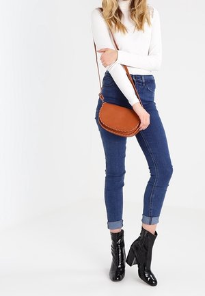 Borsa a tracolla in pelle color cuoio con dettagli intrecciati, abbinata a jeans skinny blu e stivaletti neri alla caviglia. Presenta una tracolla.