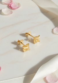 Guldiga geometriska stiftörhängen med en vågig design. Slät yta. Mot en ljus marmorbakgrund med rosa kronblad och band.