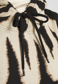 Bluza z kapturem w kolorze beżowym z czarnymi i brązowymi abstrakcyjnymi paskami. Posiada czarne sznurki z metalowymi końcówkami. Miękka tekstura materiału.