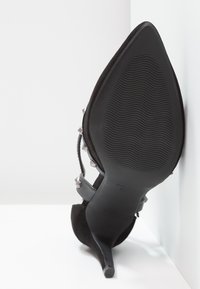 Chaussure noire en suède à talon haut avec un bout pointu, une semelle texturée et des clous en argent sur des sangles en cuir. La hauteur du talon est d'environ 10 cm.