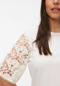 Witte t-shirt met bloemenkantdetail op de korte mouwen. Gladde stof met een ronde halslijn, die complexe kantpatronen op de mouwen toont.