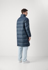Marineblauer Daunenmantel mit hohem Kragen, gestepptem Design und langem Schnitt. Kombiniert mit hellblauen Jeans und weißen Sneakers. Glatte Textur.