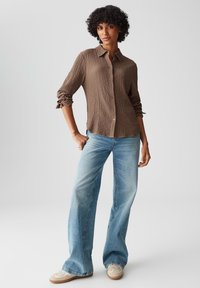 Braun strukturierte Knopfhemd mit hochgerollten Ärmeln, kombiniert mit hellblauen Schlagjeans und beigen Sneakers. Einfaches Design, lässiger Look.