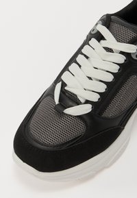 Schwarze Sneaker aus Mesh- und Wildledermaterialien, mit weißen Schnürsenkeln, abgerundeter Spitze und strukturiertem Design mit grauen Akzenten.