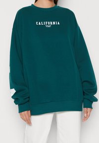 Tyrkysová mikina s kulatým výstřihem, volným střihem a bílým nápisem "CALIFORNIA". Má dlouhé, oversized rukávy a žebrované manžety.