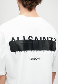 Balta kokvilnas T-kreklu ar lielu melnu grafisko apdruku uz aizmugures, uz kuras ir vārds "ALLSAINTS" un zem tā "LONDON", redzama tekstūras detaļa.