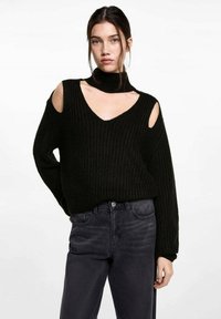 Čierny pletený sveter s výstrihom do V, s vykrojenými ramenami a vysokým golierom, v kombinácii s tmavošedými džínami. Textúrovaná tkanina s voľným strihom.