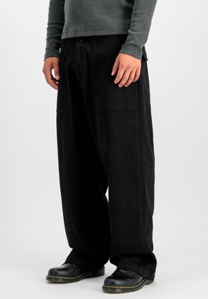 Pantaloni neri a gamba larga realizzati in tessuto resistente. Presentano grandi tasche laterali e una vestibilità comoda. Abbinati a scarpe nere con dettagli gialli.