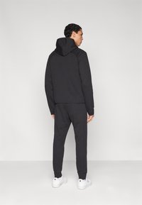 Felpa nera in cotone con cappuccio, abbinata a joggers coordinati. Tessuto liscio, design aderente e polsini a coste. Indossata con sneakers bianche.