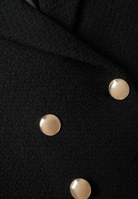 Czarna tkana tkanina płaszcza o wzorze w jodełkę, z trzema błyszczącymi złotymi guzikami umieszczonymi po lewej stronie.