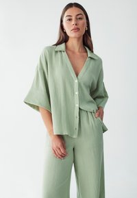 Camisa de botonadura en verde claro, texturada, con escote en forma de V y mangas cortas y anchas, combinada con pantalones sueltos a juego.