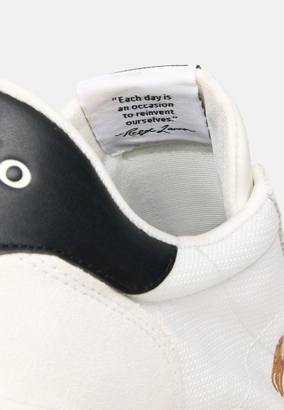 Witte sneaker met gestructureerde stof, zwart leren accent en een zichtbaar label met een citaat aan de binnenkant van de tong.
