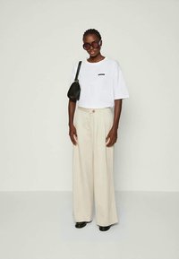 Λευκό κοντό μπλουζάκι με μικρό μαύρο λογότυπο, συνδυασμένο με κρέμα παντελόνι με φαρδιά πόδια, μαύρη τσάντα χειρός και oversized γυαλιά ηλίου. Λείες υφές.