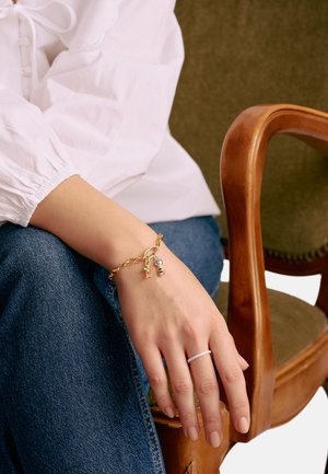 Goldene Kettenarmband mit Anhängern, darunter ein klarer Edelstein. Schlanker silberner Ring mit kleinen Steinen, getragen an einer entspannten Hand auf Denim-Stoff.