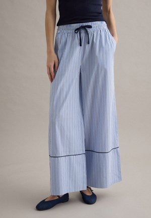 Frau trägt weit geschnittene blau-weiße vertikal gestreifte Hosen mit Kordelzugbund und dunkelblaue Ballerinas, steht mit Händen in den Taschen.