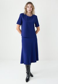 Ensemble deux pièces bleu marine comprenant un haut à manches courtes et une jupe fluide. Tissu lisse avec une finition élégante. Bottes noires montant jusqu'au genou.