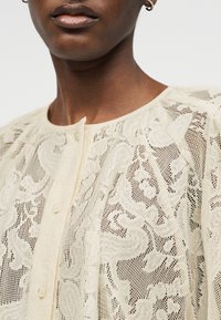 Κοντινό πλάνο ενός ατόμου που φορεί κορυφή από κρεμ δαντέλα με λουλουδάτα σχέδια, κουμπωμένη στο λαιμό, και μικρά σκουλαρίκια κρίκοι.