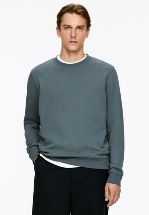 Gray cotton sweatshirt with a crew neck, long sleeves, and ribbed cuffs. Worn over a white t-shirt, paired with dark pants.