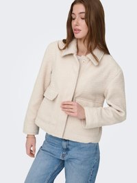 Veste matelassée de couleur crème avec col, poches avant et fermeture à boutons-pression. Le tissu a une finition texturée. Portée avec un jean bleu clair.