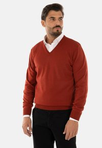 Maglione rosso a collo a V realizzato in un tessuto knit liscio, con polsini e orlo a costine. Indossato sopra una camicia bianca con colletto, abbinato a pantaloni neri.