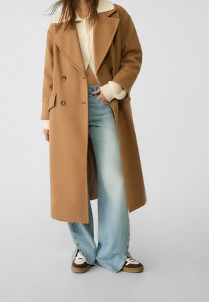 Personne portant un long manteau camel par-dessus un pull crème, un jean large bleu clair, et des baskets marron et blanches sur un fond clair.