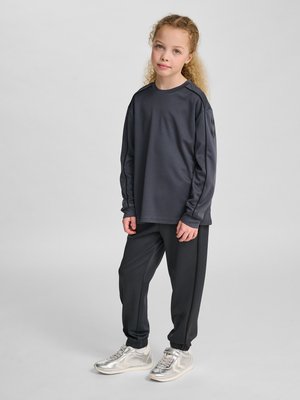 Młoda dziewczyna z kręconymi włosami, ubrana w ciemny top z długim rękawem, dopasowane spodnie dresowe i srebrne trampki, stoi na jednolitym tle.