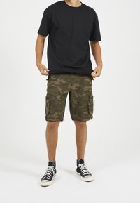 Uomo che indossa una maglietta nera, pantaloni cargo mimetici verdi e scarpe nere alte, in piedi contro uno sfondo bianco.