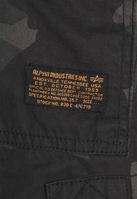 Black fabric with a camouflage pattern; features a rectangular patch with orange text, branded "Alpha Industries Inc." and manufacturing details.