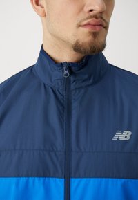 Giacca zip-up blu navy con colletto alto e tessuto testurizzato. Presenta un logo grigio sul petto e un dettaglio della cerniera liscio.