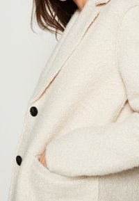 Abrigo de textura crema con cuello en pico, botones negros y bolsillos laterales. La tela tiene un aspecto suave y afelpado.