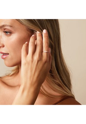 Gold ring with two white pearls and small gold accents, worn on a hand with light skin and natural nails, against a soft beige background.