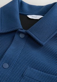 Cămașă din material albastru cu textură tip nervuri, cu guler clasic, nasturi capsa, buzunar frontal și etichetă albă vizibilă în interiorul gulerului cu inscripția "NEXT".