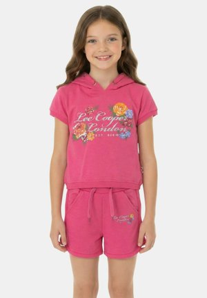 Fille souriante portant un sweat à capuche rose à fleurs et un short assorti avec le texte « Lee Cooper London », debout devant un fond blanc.