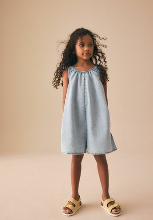 Młoda dziewczyna z długimi kręconymi włosami, ubrana w jasnoniebieski bezrękawnik z denimu i żółte sandały, stojąca na beżowym tle.