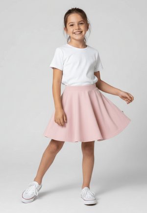 Uśmiechnięta dziewczyna w białej koszulce, różowej spódnicy i białych trampkach, stojąca z jedną nogą wyciągniętą do przodu i trzymająca spódnicę.