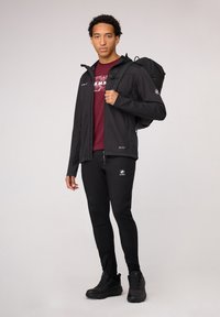 Zwarte softshell jas met ritszakken, wijnrood T-shirt met grafisch ontwerp, zwarte strakke broek en zwarte wandelschoenen, staande tegen een lichte achtergrond.