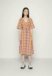 Mulher a posar vestindo um vestido midi de xadrez colorido com botões à frente e ténis brancos robustos, num fundo simples.