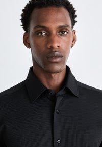 Camisa negra de botón con un tejido texturado, que presenta un cuello estándar y botones en contraste. Diseño ajustado, sin patrones visibles.