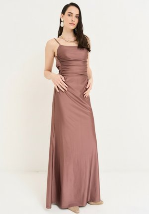 Kvinde i en mauve satin spaghetti strop kjole, poserer med hænderne på hofterne, iført markante øreringe og skalhalskæde, mod en ensfarvet baggrund.